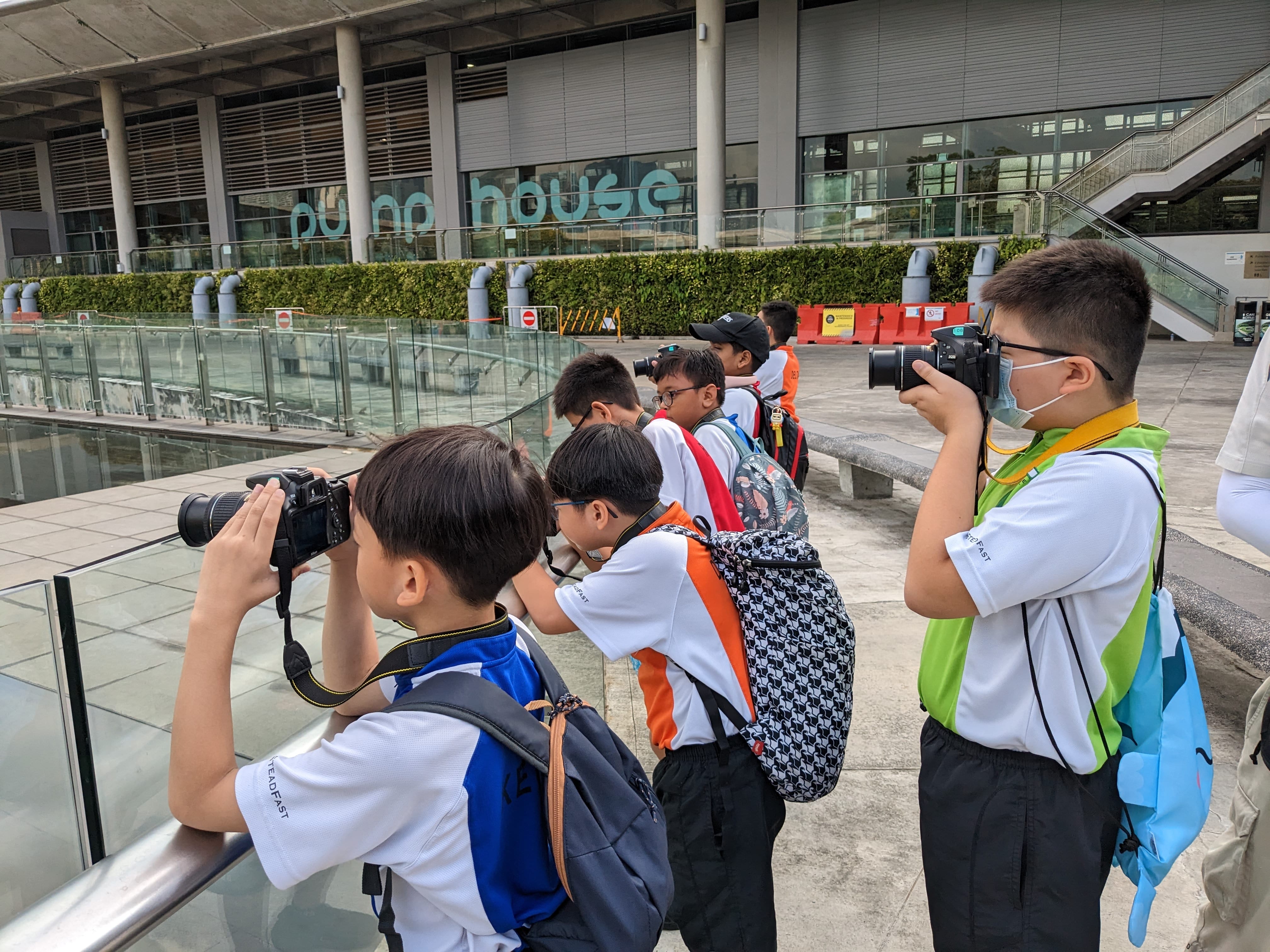 Several young students with backpacks hold DSLR cameras outdoors in front of a building labeled Pump House.